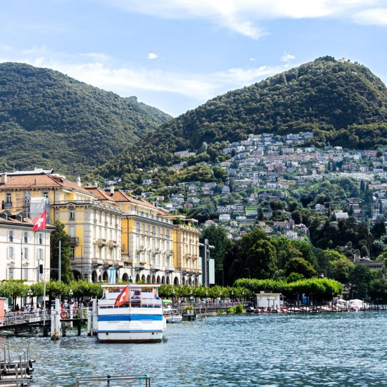 Scenic view of Lake Lugano with mountains and elegant buildings against a bright sky.