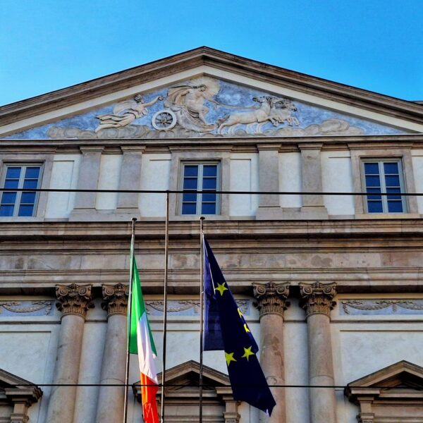 Front view of La Scala opera house facade with Italian and EU flags under blue sky.